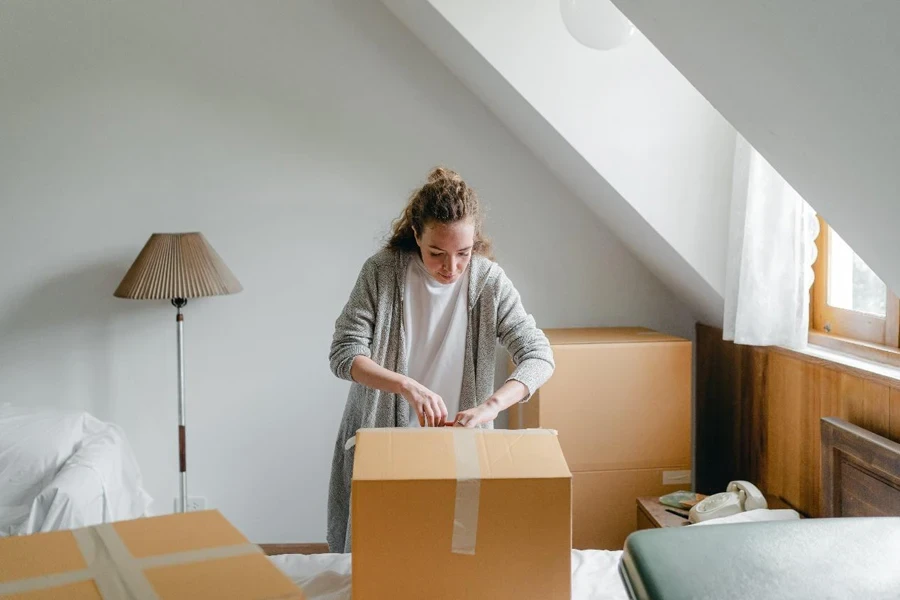 Slim female in cardigan unpacking cardboard box put on bed close to suitcase while standing near window in attic style room in daylight