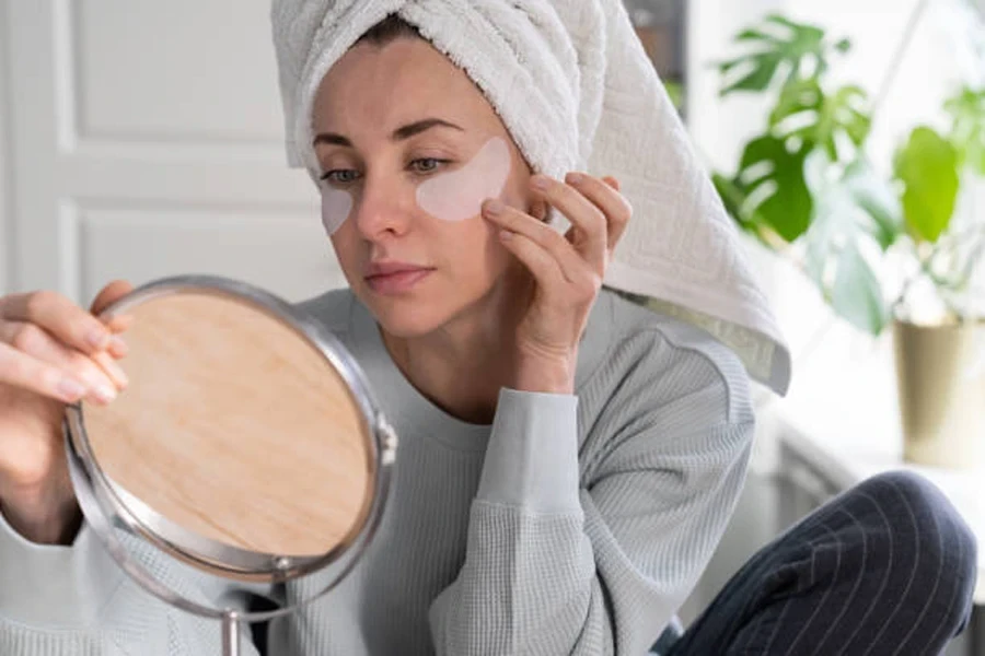 Une femme avec une serviette sur la tête applique des patchs hydrogels pour le contour des yeux à la maison