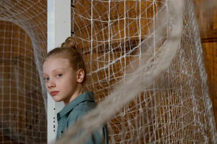 Girl in Gray Shirt Looking at Camera While Standing Beside the Climbing Net