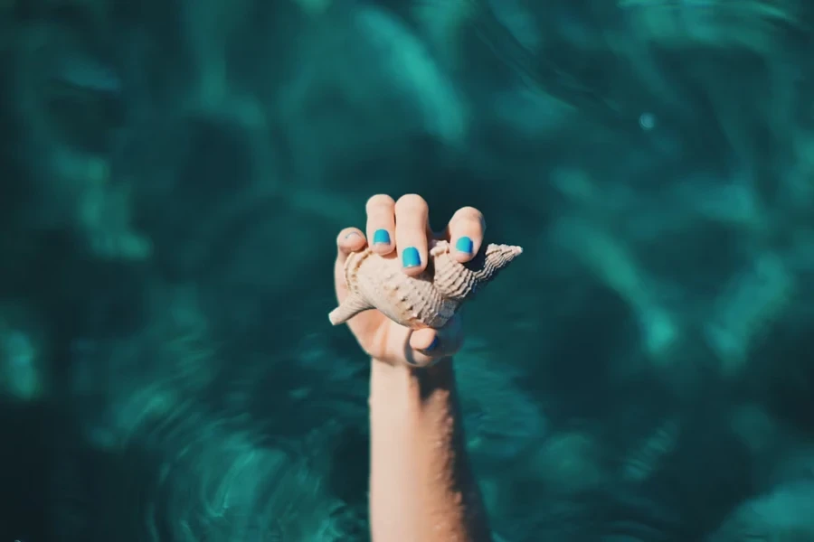 Hand mit blauem Nagellack hält Muschel im Wasser