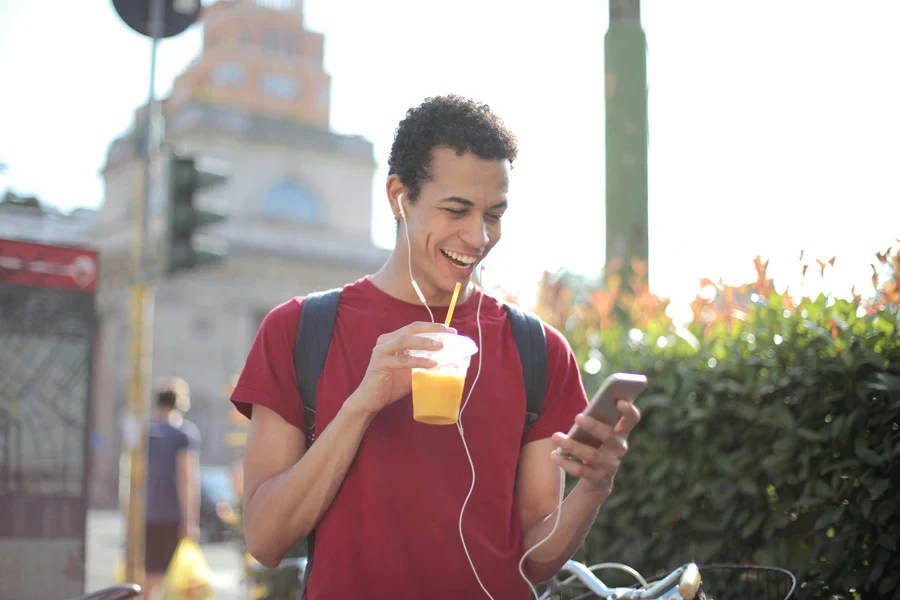 Cara étnico feliz em traje casual e com mochila tomando um copo de suco fresco usando smartphone enquanto está na rua e ouve música