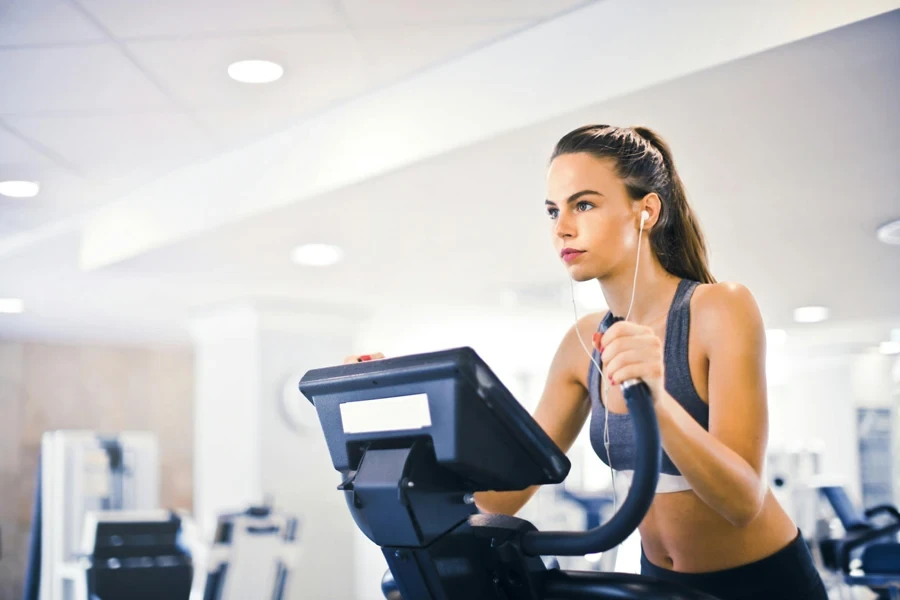 Mulher séria em fones de ouvido e roupas esportivas ouvindo música e correndo na esteira no centro esportivo contemporâneo leve