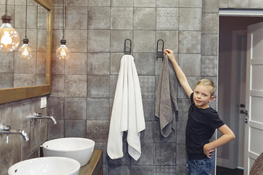 Niño pequeño con cabello rubio colgando una toalla en un baño espacioso mientras mira a la cámara