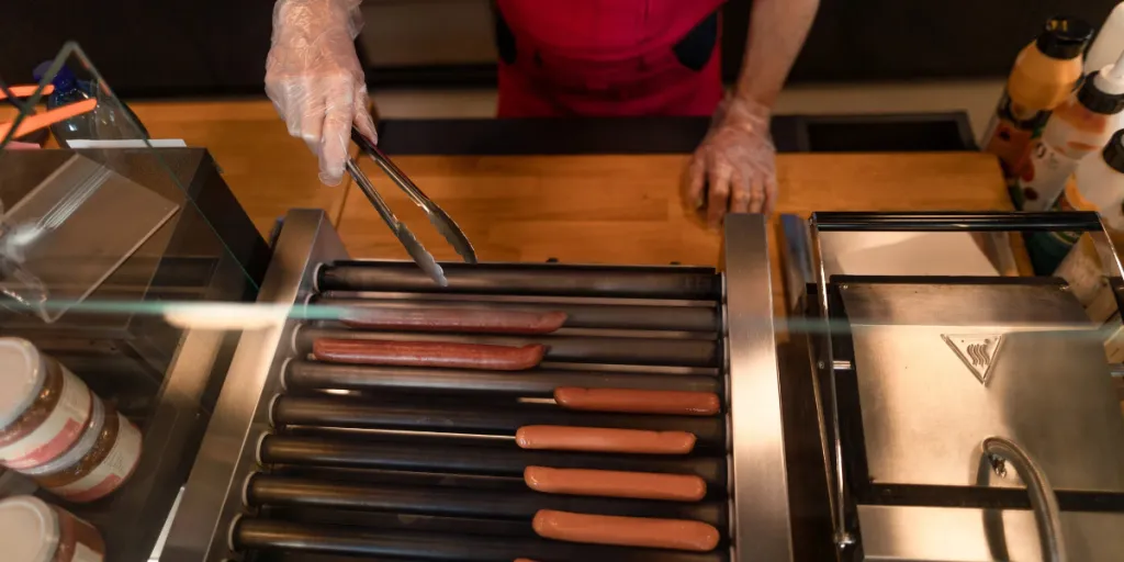 A close-up of waiter standing by counter and prepairing hot-dog to a customer in cafe at gas station