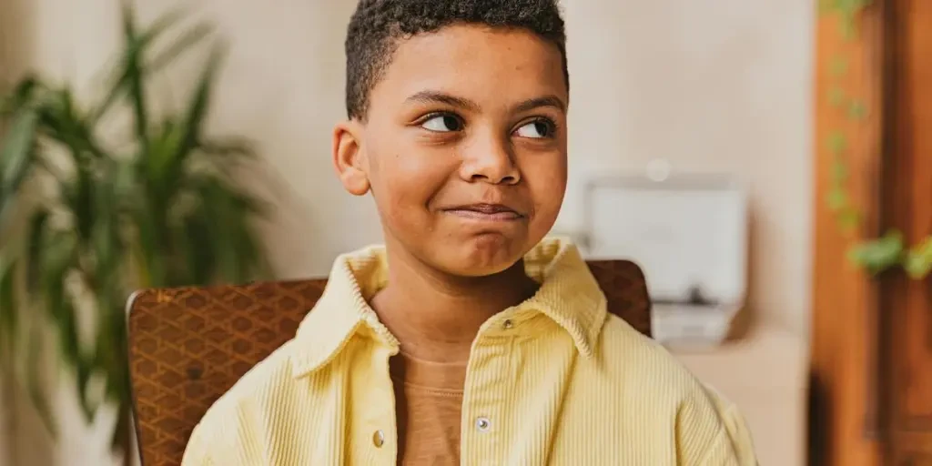 A happy child with curly hair smiles mischievously, wearing a yellow shirt indoors