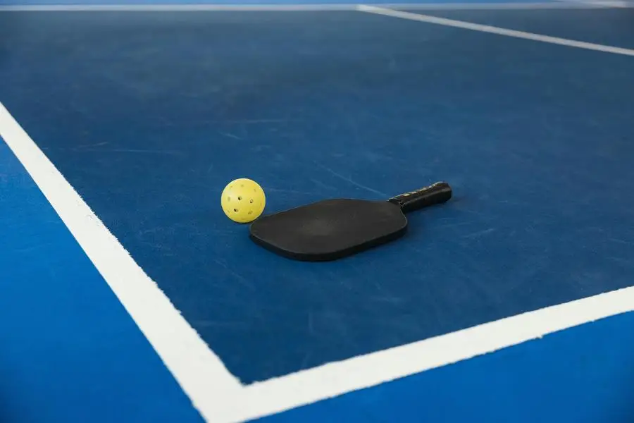 Una paleta de pickleball y una pelota amarilla descansando sobre una cancha de pickleball azul con líneas blancas