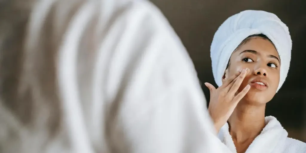 A young woman applies skincare cream while wearing a bathrobe in front of a mirror