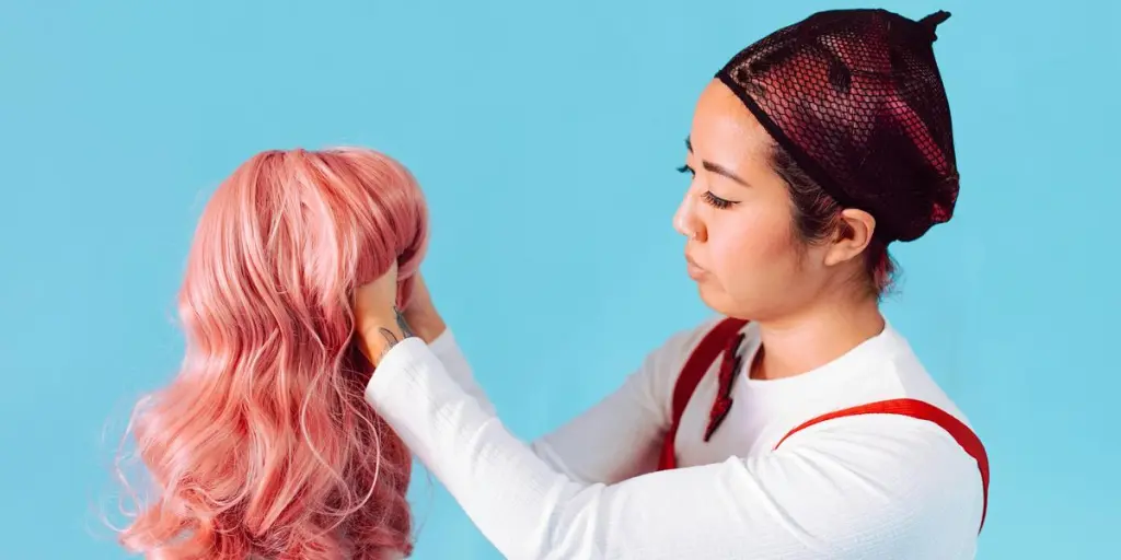 An Asian woman in a net cap holds a pink wig against a blue backdrop, showcasing style and hair fashion