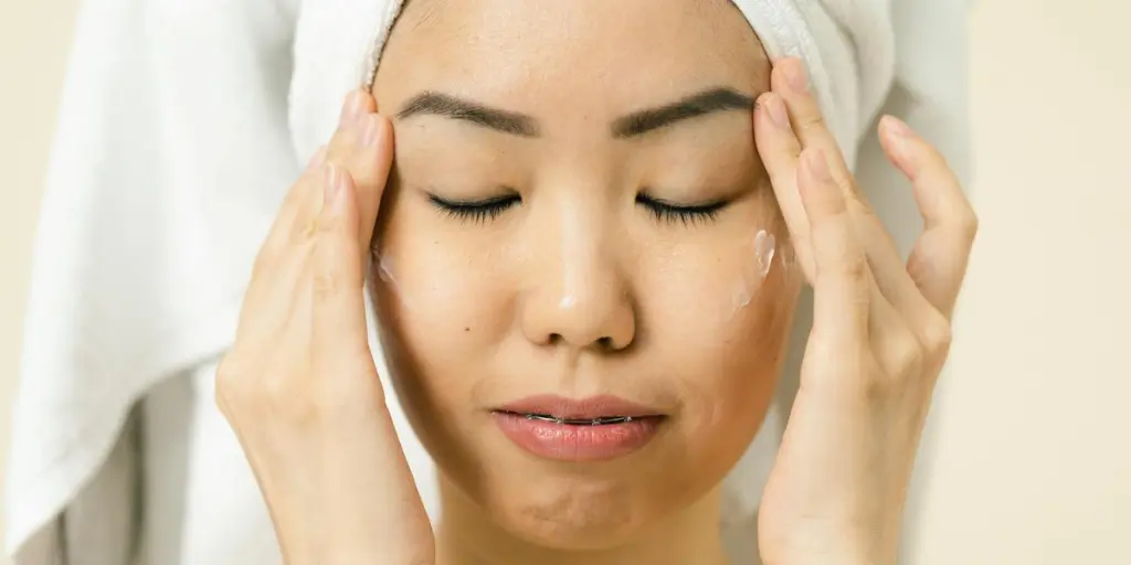 Asian woman with a towel on head applying moisturizer for skincare routine