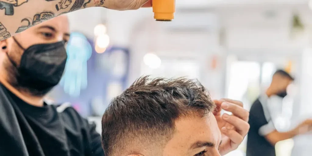 Barber throwing talcum powder on the hair of a caucasian man wearing a mask in a barbershop