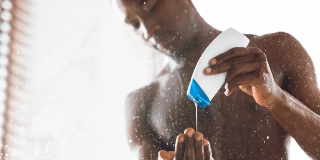 Hombre negro vertiéndose gel de ducha mientras se lava el cuerpo desnudo bajo el agua que cae en un baño moderno en casa