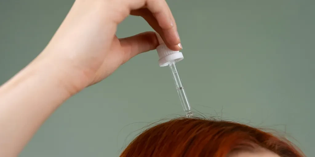 Gros plan d'une main appliquant du sérum capillaire sur des cheveux roux à l'aide d'un compte-gouttes