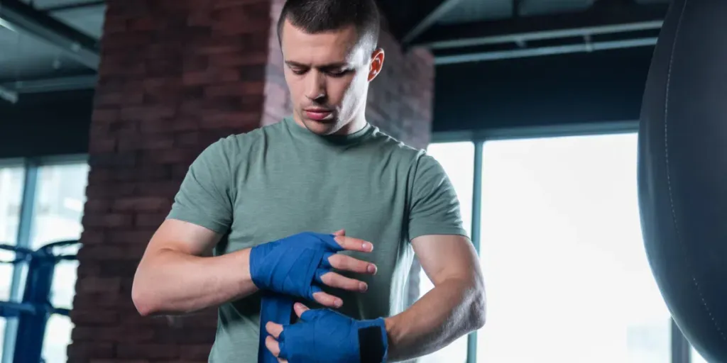 Dark-haired promising skillful boxer using blue wrist wraps in light modern gym