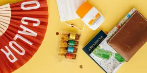 Flatlay of travel essentials like mask, vaccine, and sunscreen on yellow background