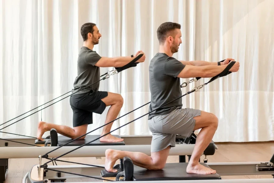 Full body side view of strong men in sportswear doing hug tree kneeling pilates exercise with resistance bands on reformer bed during training in gym