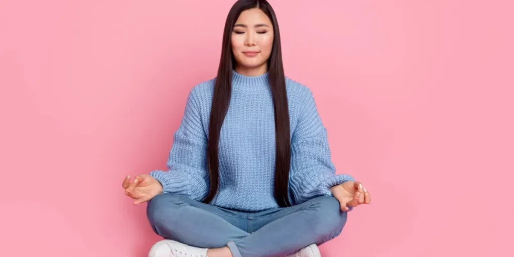 Photo pleine longueur d'une femme brune millénaire endormie assise en train de méditer, portant un pull, des jeans et des chaussures isolées sur fond de couleur rose