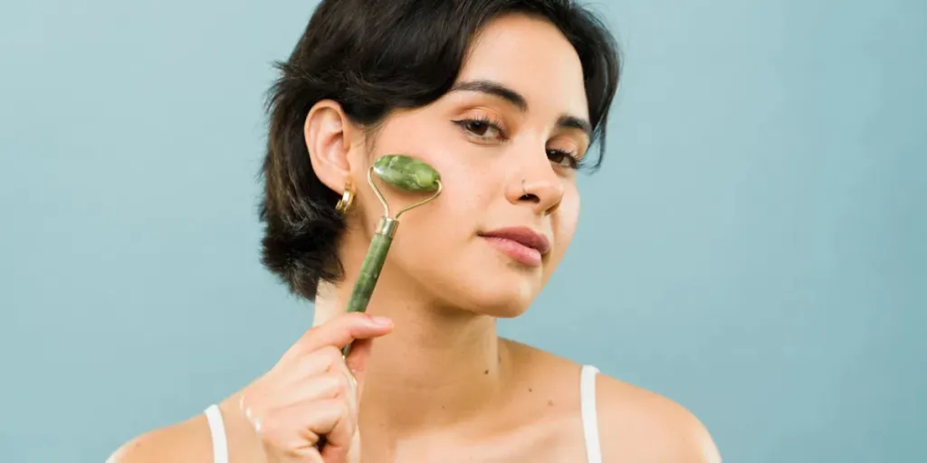 Gorgeous woman using a jade roller for facial massage in a studio with a blue backdrop for skincare routine