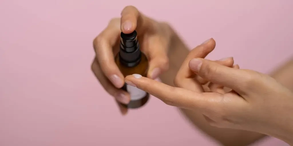 Hands applying skincare serum from a spray bottle against a pink background