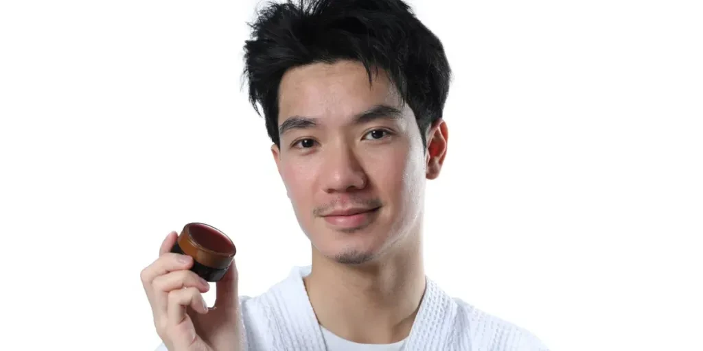 Hombre guapo sosteniendo un frasco con pomada o cera para peinar el cabello sobre fondo blanco