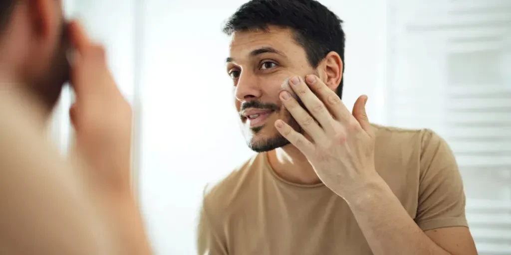 Handsome man standing in the bathroom and looking at the mirror while applying face cream or anti aging moisturizer