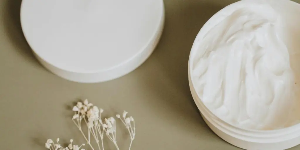 Natural skincare cream in white jar with delicate flowers on green background