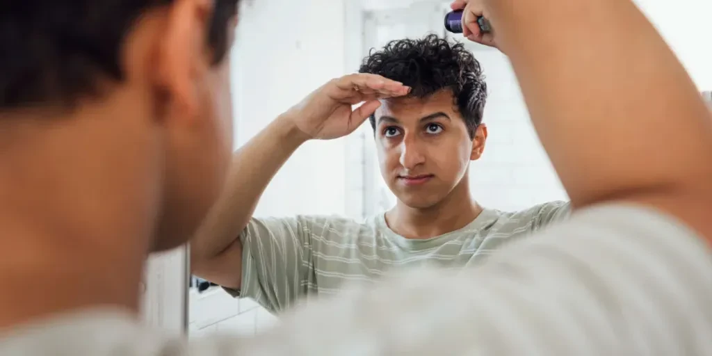 Vista por encima del hombro de un joven con cabello rizado parado en su baño en una mañana.