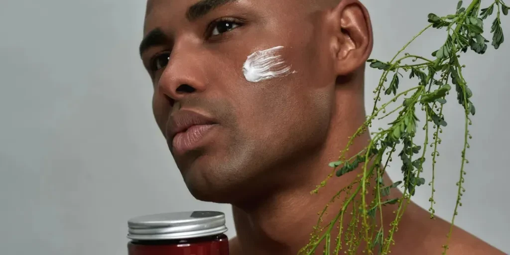 Person demonstrating his cheek with mark of cream taking hold of a red jar and small branch