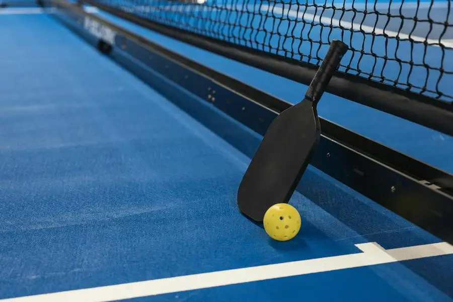 Paleta de pickleball apoyada contra una red con una pelota amarilla en una cancha azul