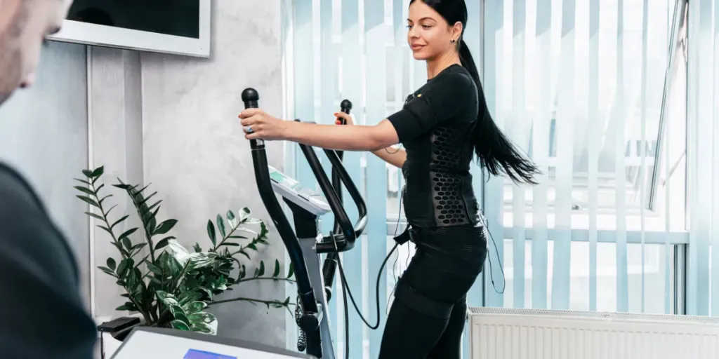 Professional physiotherapist working with beautiful young woman in EMS suit during special electro stimulation training on a stepper or cardio climber