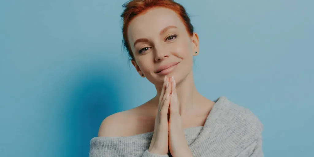 Femme rousse sereine avec les mains jointes, pull à épaules dénudées, sur fond bleu