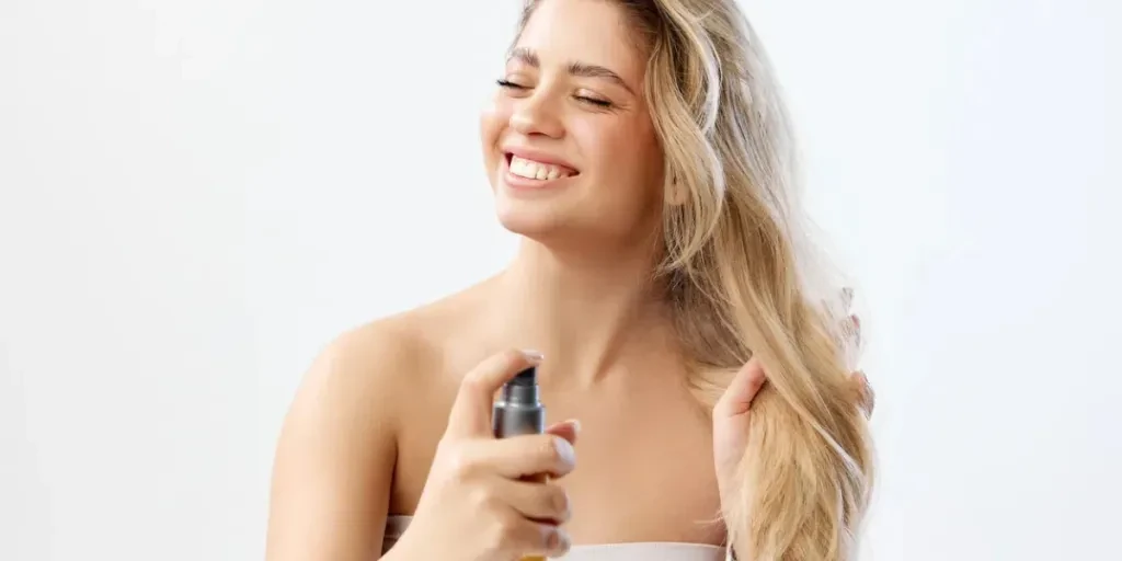 Mujer sonriente con top sin tirantes y cabello rubio largo aplicando spray contra un fondo de estudio blanco