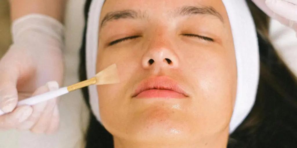 Vue de dessus d'une cosmétologue qualifiée en uniforme et gants appliquant de l'huile essentielle sur la peau du visage d'une jeune femme tout en travaillant dans un centre de spa contemporain