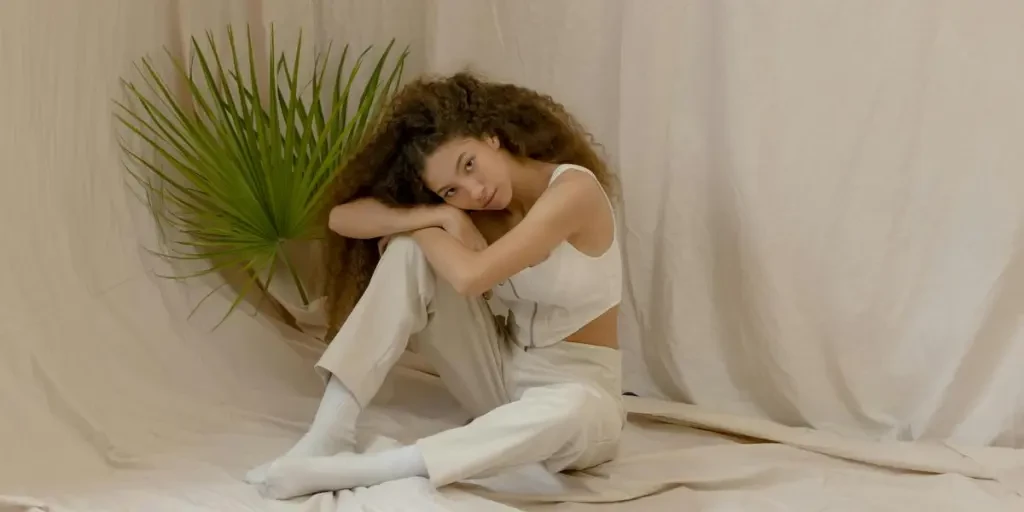 Femme aux cheveux bouclés dans une tenue blanche posant sur un fond neutre avec des feuilles de palmier