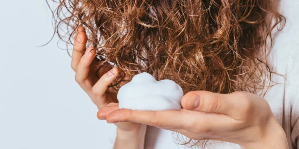 Woman's hands apply styling mousse to her curly hair on white studio background, close-up