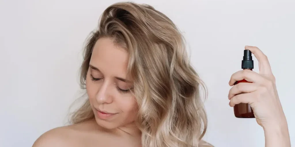 Joven caucásica atractiva mujer rubia con cabello ondulado rociando laca de protección térmica