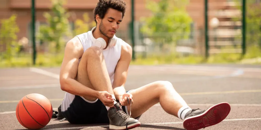 Atleta joven de raza mixta con ropa deportiva sentado en una cancha de baloncesto y atándose los cordones de las zapatillas antes del entrenamiento