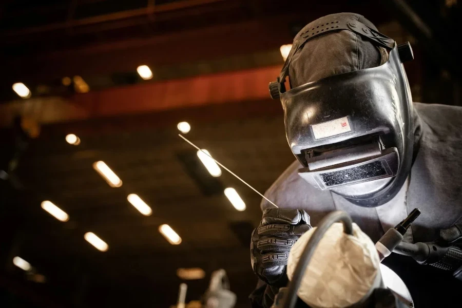 A man using a Heliarc welder, showing the gas tube, metal filler rod (TIG rod), and electrode torch