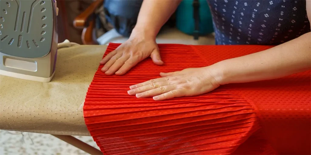 lose-up of hands smoothing red pleated fabric on an ironing board next to a steam iron