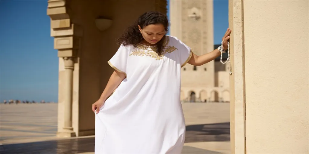 young woman wearing a white embroidered dress stands thoughtfully in front of a mosque on a sunny day