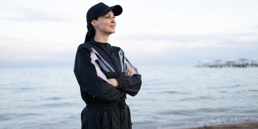 Eine schöne Brünette Frau trägt Mode Sportanzug und Mütze Blick auf den Sonnenuntergang am Strand