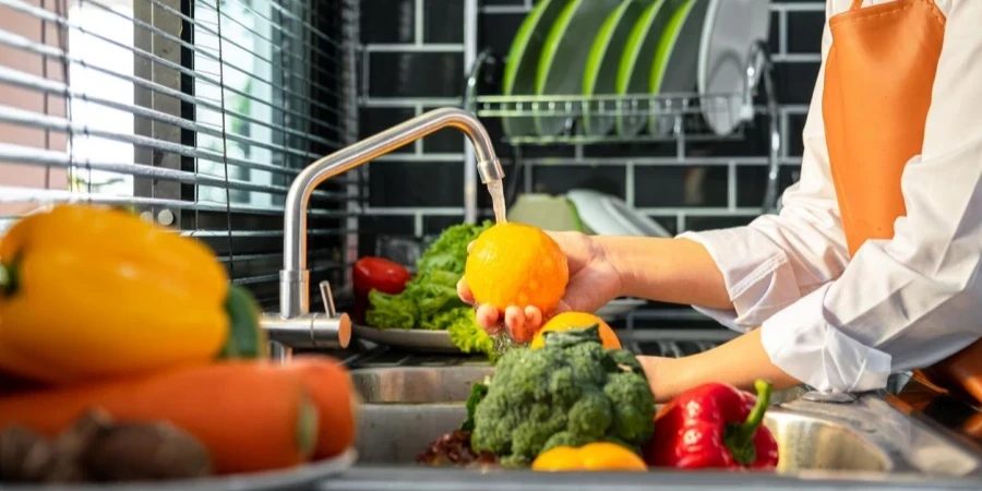 Mãos de dona de casa asiática lavando laranja e vegetais com água na pia enquanto usa avental