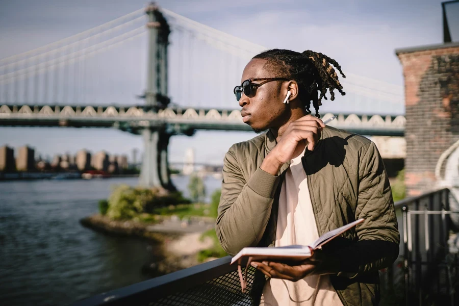 Wistful African American guy in wireless earphones and sunglasses standing with open organizer and pen leaned on bridge fence while looking at river in daylight and listening to music