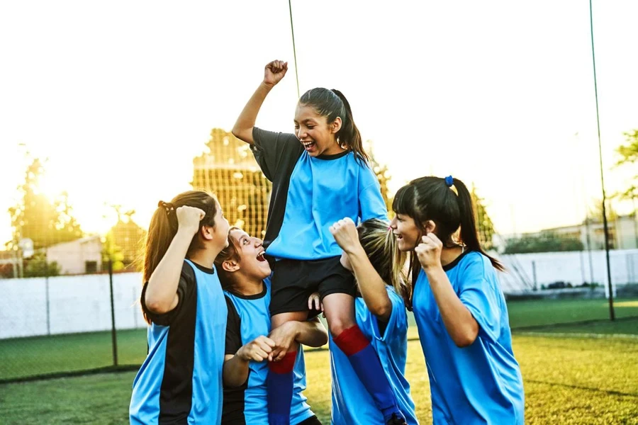 Vrolijke meisjesvoetballer die een teamgenoot op hun schouders dragen na het winnen van een wedstrijd. Het vrouwelijke voetbalteam viert hun succes en geniet van momenten samen op het buitenveld.