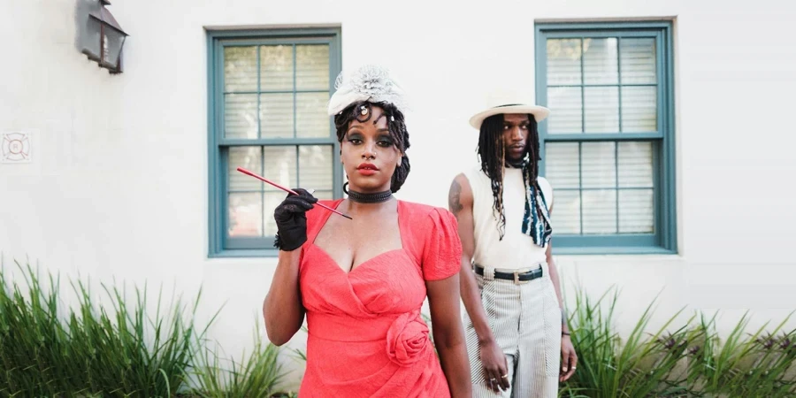 Une femme vêtue d'une élégante robe rouge tenant des cigarillos et un homme avec des dreadlocks debout derrière