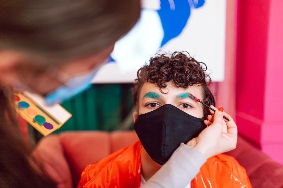 Man in a face mask getting his eyebrow colored teal