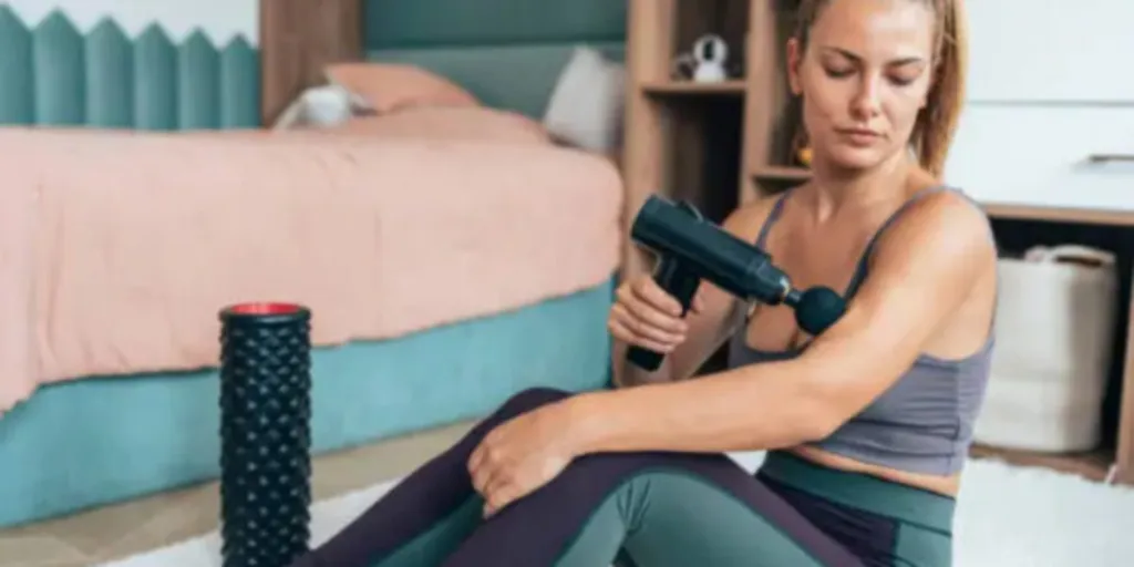 Woman sitting next to bed using massage gun on arm