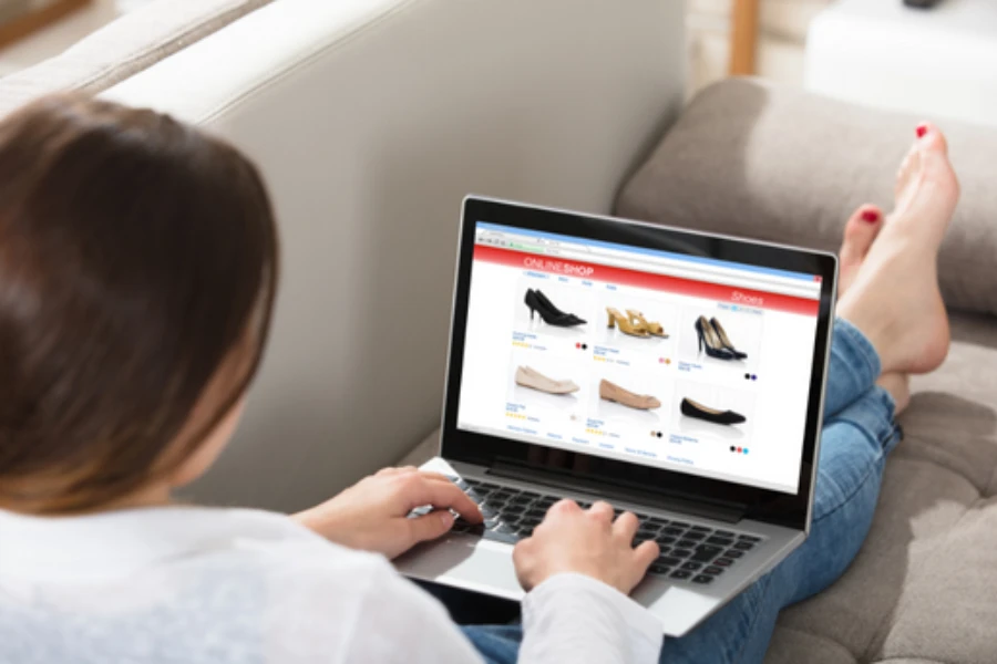 女性がソファに寝そべり、膝の上にコンピューターを置き、靴のショッピング サイトを表示している