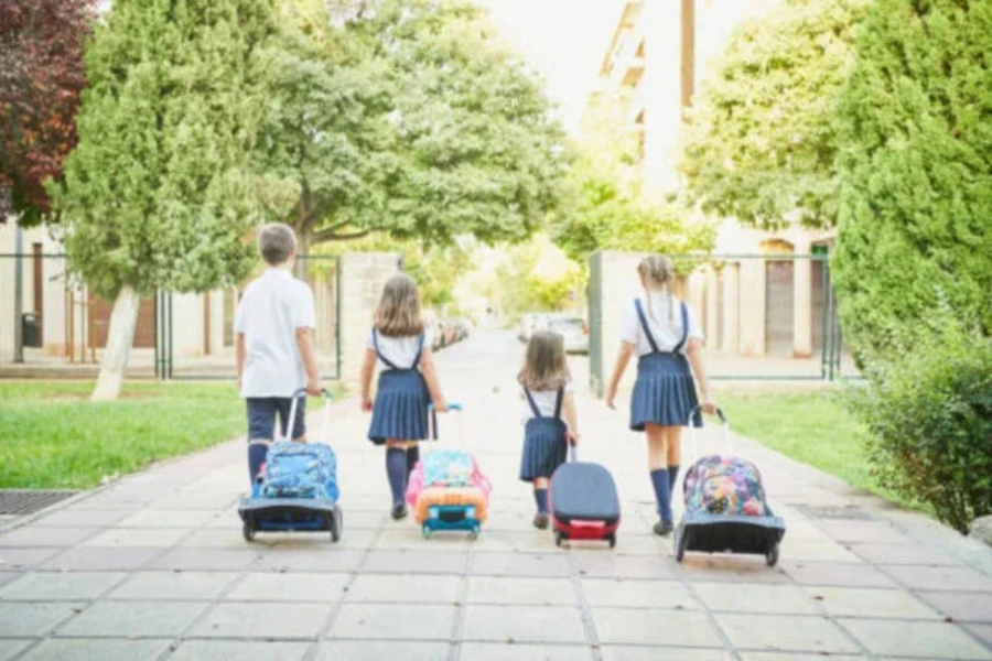 Four kids wheeling backpacks through a park area