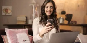 Woman sitting on sofa while looking at phone with laptop on lap