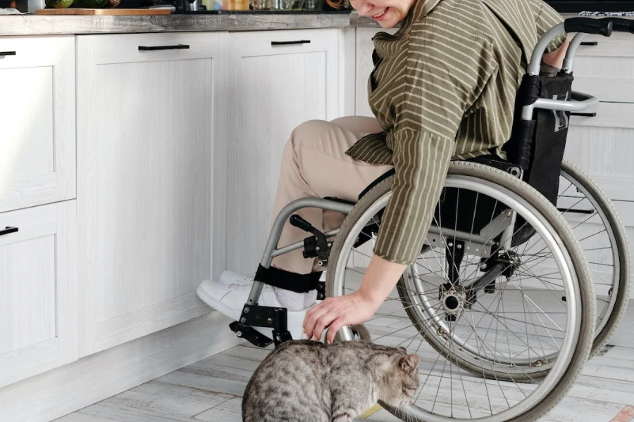 Person in a wheelchair in the kitchen leaning over to pet a cat